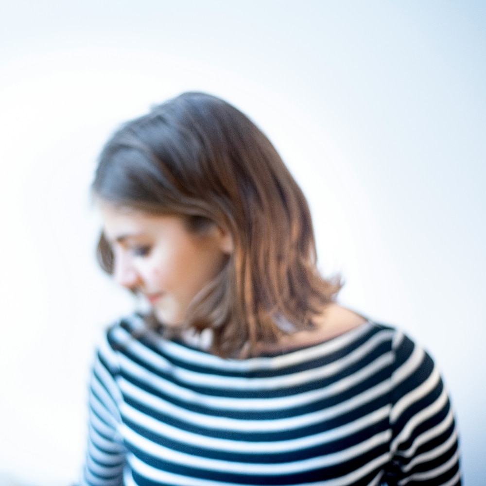 A woman with shoulder-length brown hair, wearing a black and white striped shirt, looks down with her face slightly out of focus against a light background.