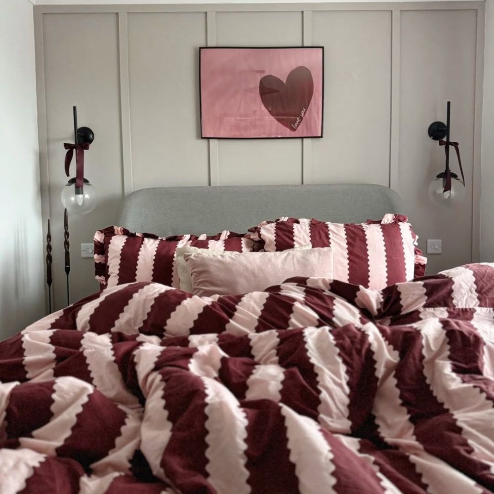 A neatly made bed with maroon and pink zigzag bedding invites you to rest in front of a gray headboard. Above the bed, a framed pink heart artwork hangs while two wall-mounted lamps with black ribbons help you stay cozy.