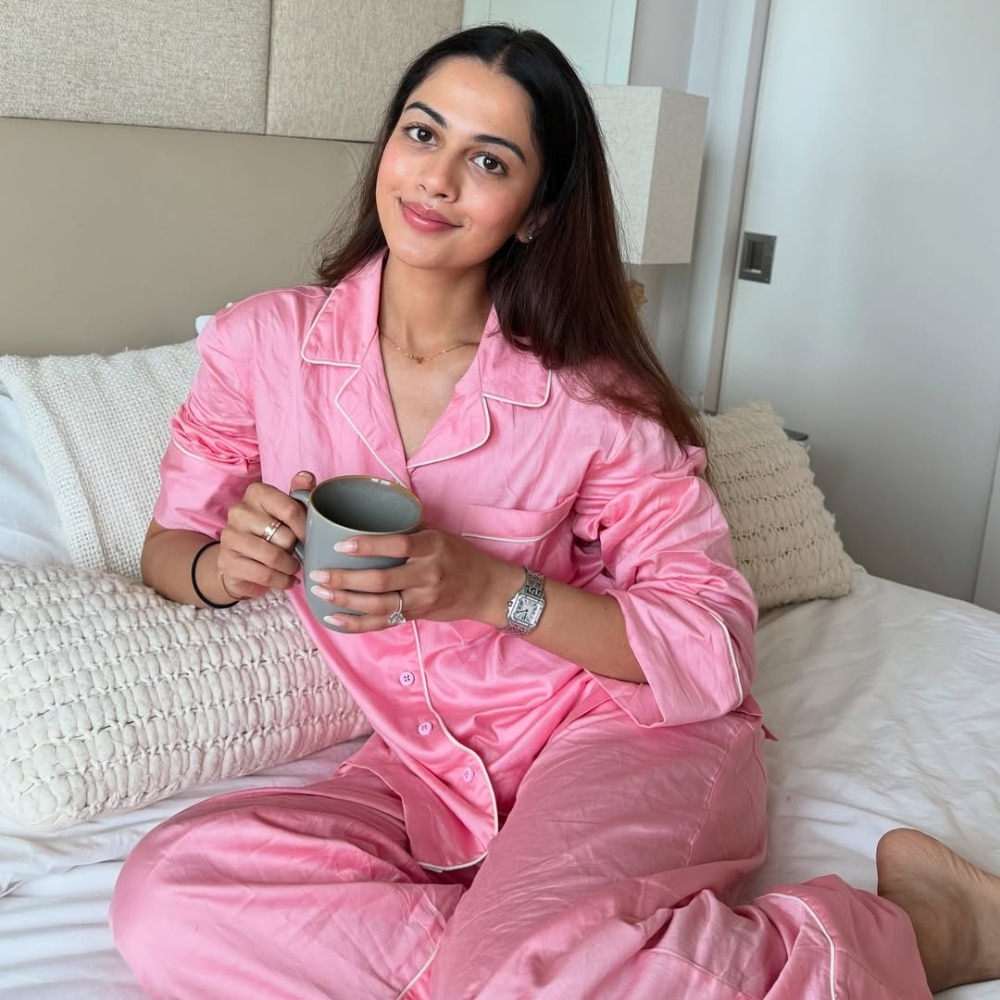 A woman in pink satin pajamas sits on a bed, enjoying a restful reset with a gray mug in hand. She smiles softly at the camera, surrounded by white bedding and pillows in her cozy, well-lit bedroom.