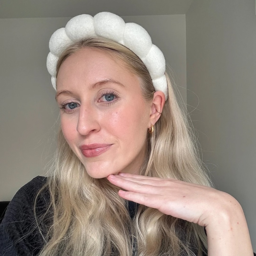 A woman with long blonde hair wearing a white, puffy headband smiles slightly and rests her chin on her hand. She is dressed in a dark top, enjoying a quiet rest indoors against a plain wall—a perfect moment for a gentle reset.