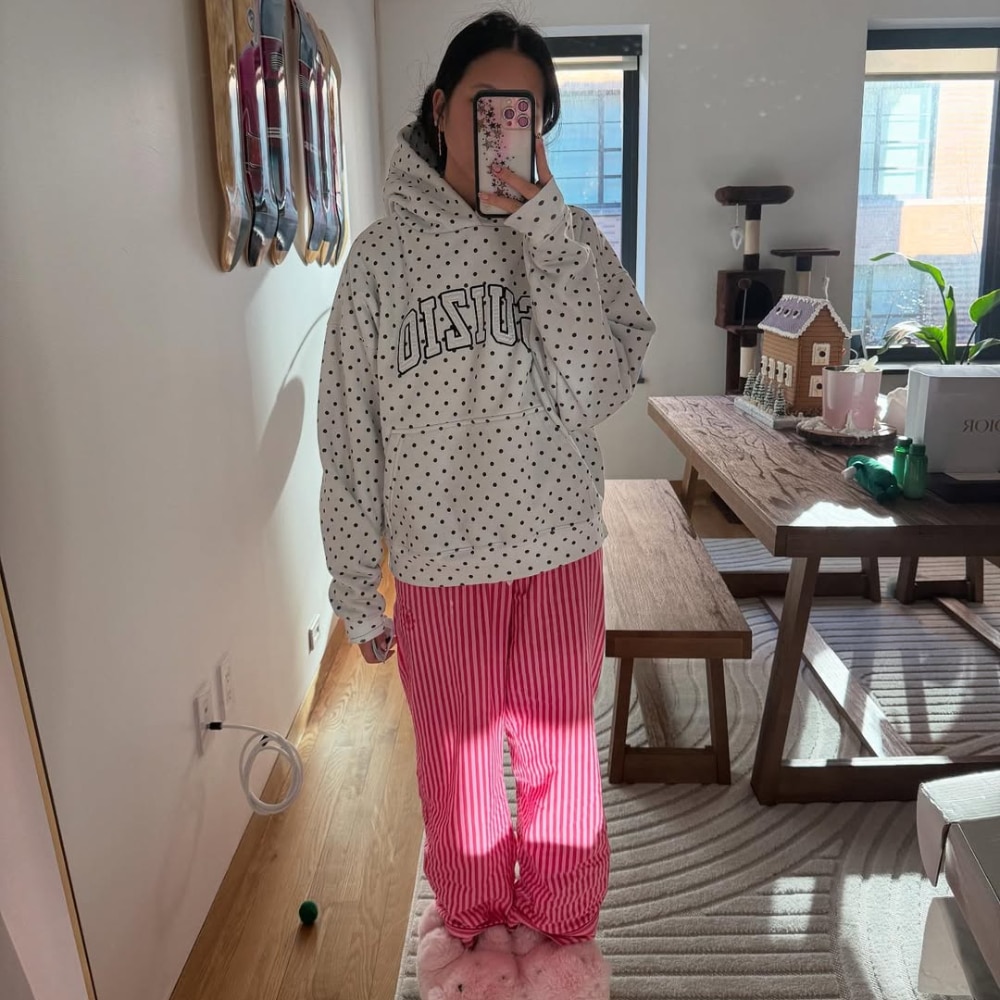 A person stands indoors taking a mirror selfie. Dressed for a rest day, they wear a white hoodie with black polka dots and BOSTON written on it, and pink striped pajama pants. Sunlight streams into the tidy room, adding warmth.