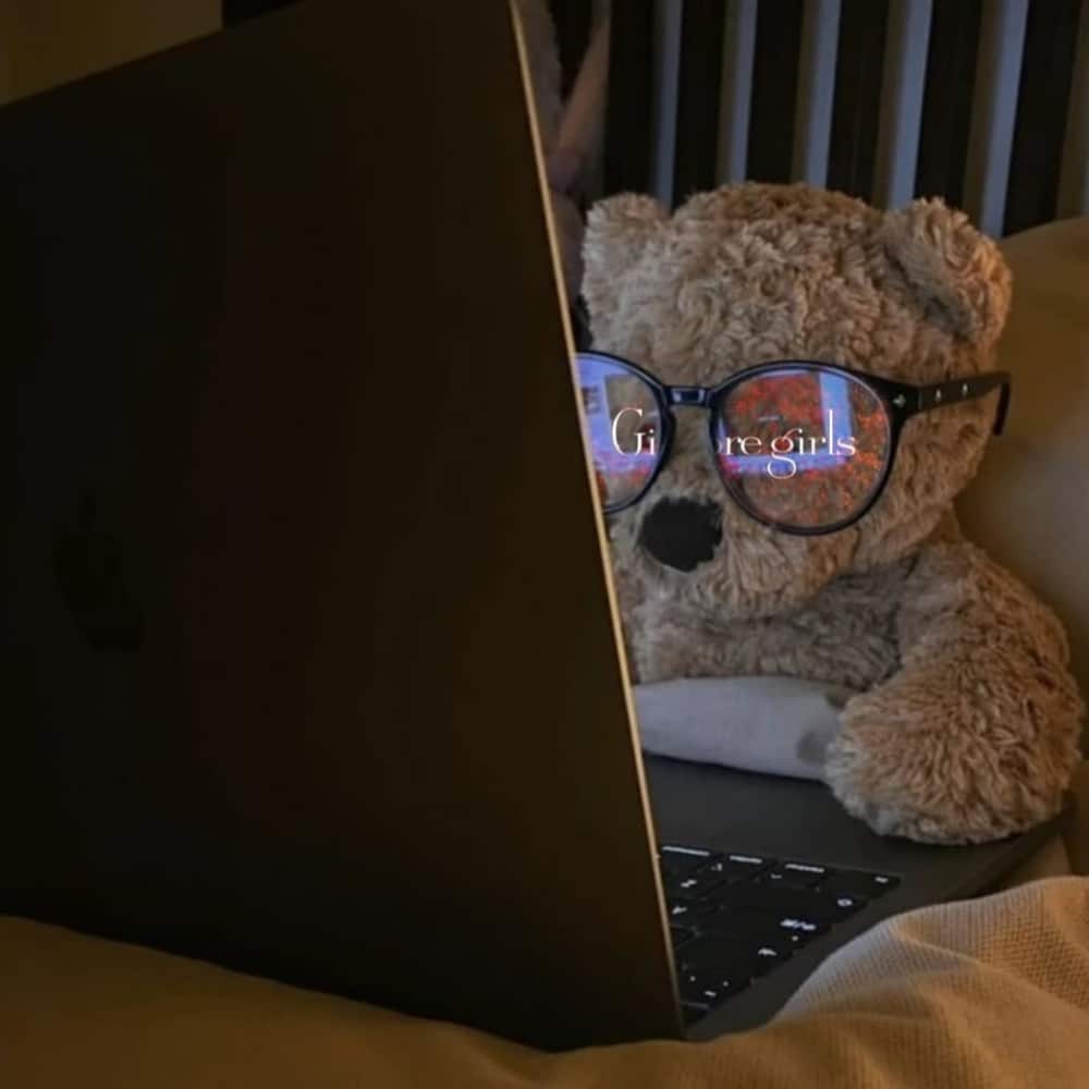 A teddy bear wearing glasses sits in bed, facing an open laptop. The screen’s reflection in the glasses shows the words “Gilmore girls.” It’s a cozy, dimly lit scene—perfect for a restful sick day and a gentle reset.