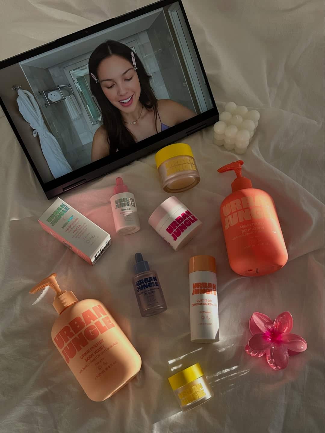 A laptop shows a smiling woman on a video call. In front of the laptop, various skincare products—including Urban Jungle bottles and jars—suggest the start of an effective skincare routine, arranged on white fabric with a pink flower and cube candle.