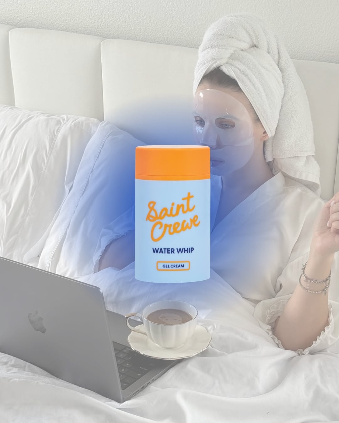 A woman in a towel robe and face mask sits in bed with a laptop and coffee, enjoying her skincare routine. In front of her is a glowing jar labeled Saint Creve Water Whip Gel Cream—a must-have among skincare products.