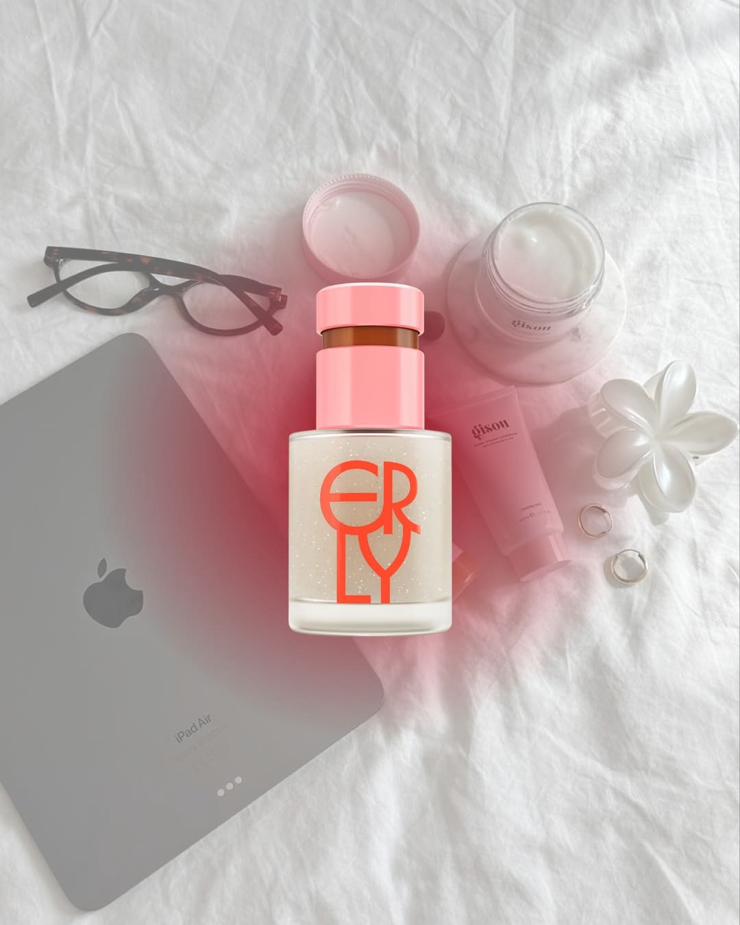 A bottle labeled ERLY sits on a white bedsheet with eyeglasses, an iPad, face cream jars, rings, and a white flower, evoking the serenity of a gentle morning skincare routine.