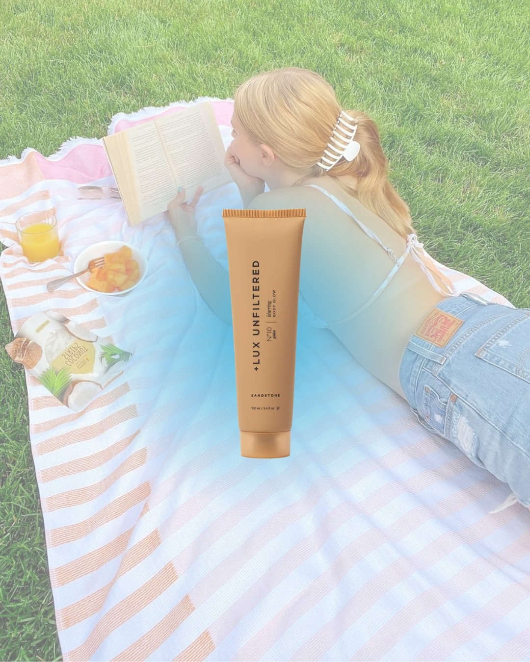 A woman practices self care on a striped picnic blanket, reading a book with a drink and fruit nearby. A large skincare lotion bottle floats centered in the foreground, highlighting her relaxing day on the grass.