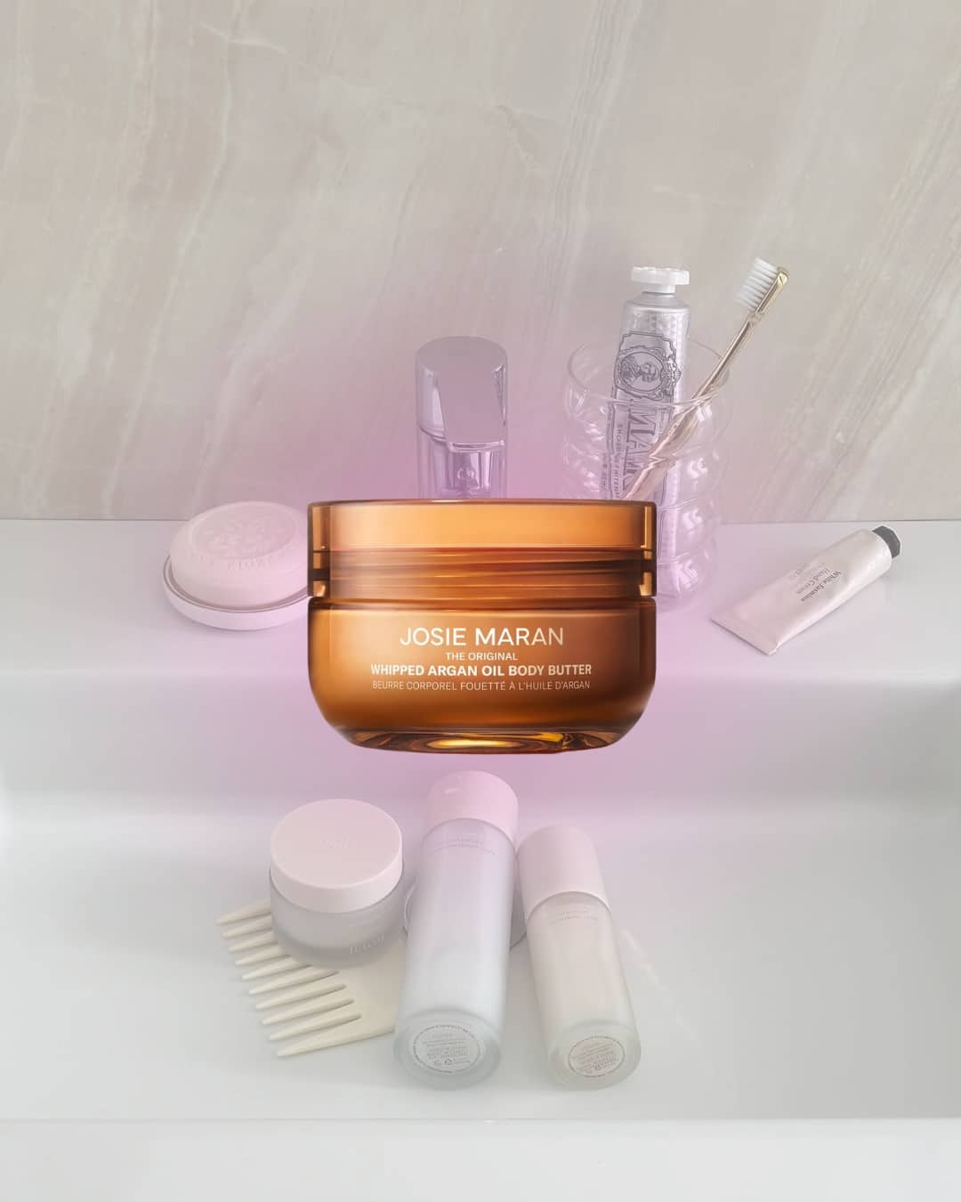 Amber jar of Josie Maran Whipped Argan Oil Body Butter on a white bathroom countertop surrounded by toiletries and skincare products, perfect for enhancing your daily skincare routine. The background features a light marble wall.