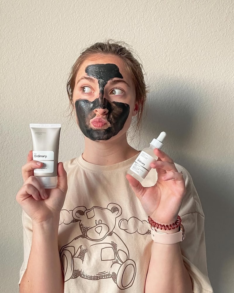 A woman with a black face mask holds up two The Ordinary products—one in a tube, one in a dropper bottle—while making a playful pout, enjoying the best at-home spa day in her beige shirt featuring a bear illustration.