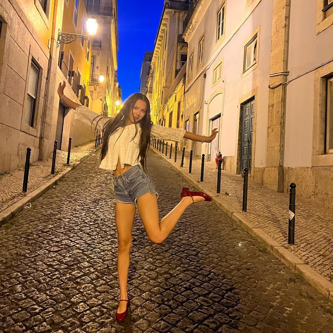 A young woman poses playfully on a cobblestone street at night, balancing on one foot with arms outstretched. She wears a white top, denim shorts, and red shoes. The street is lined with old buildings under a deep blue sky.