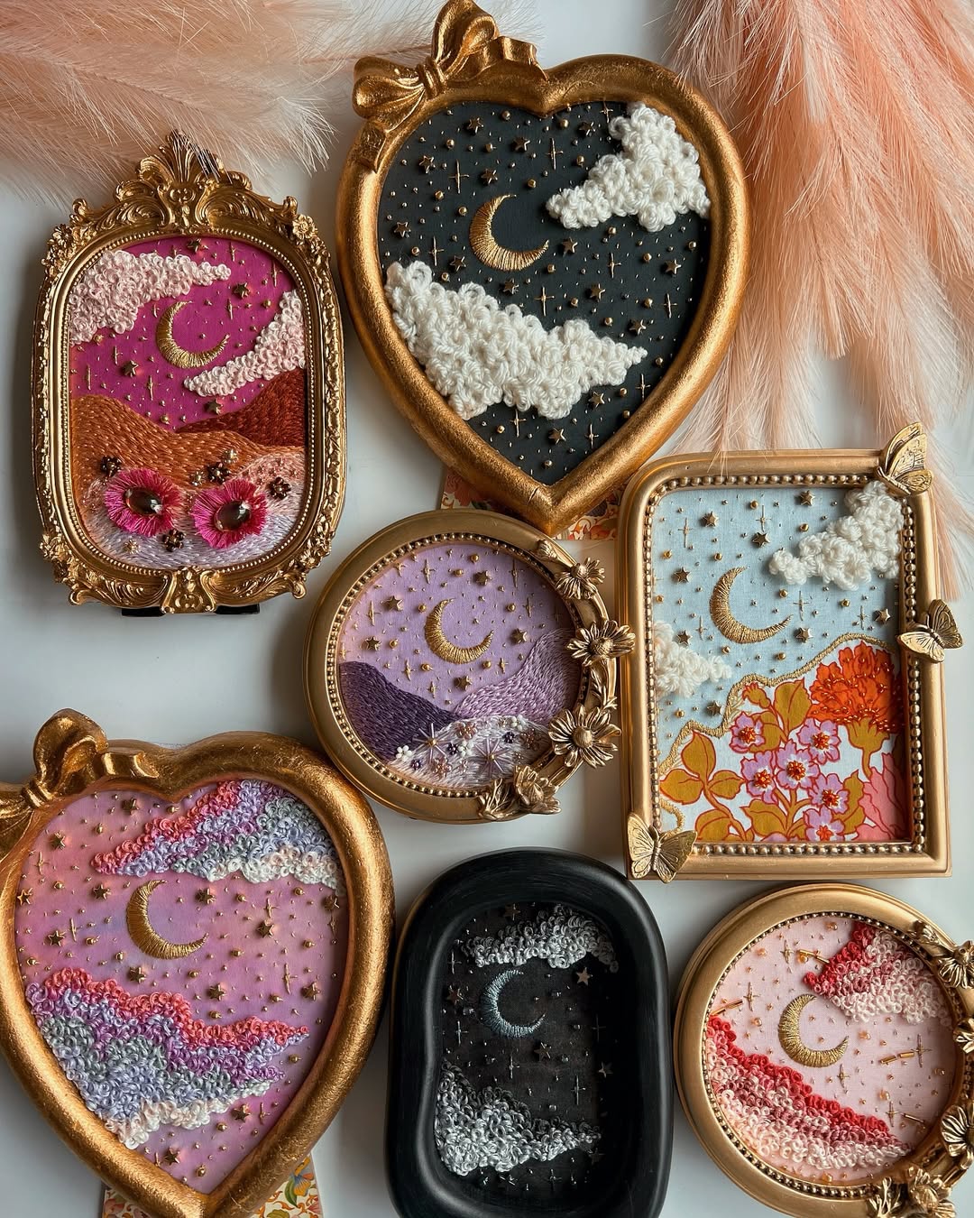 A collection of ornate, gold-framed embroidered art pieces, featuring crescent moons, clouds, and starry skies in vibrant colors and various shapes, arranged on a surface with soft peach feathers nearby.