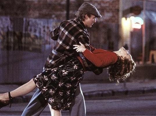 A man in a hat and plaid jacket dips a smiling woman in a red sweater and floral skirt as they dance together on a street at night. The background is softly lit with blurred buildings and lights.