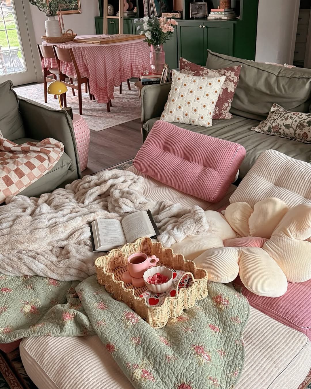 A cozy living room setup with sofas, patterned pillows, blankets, a new book, and a wicker tray holding a pink tea set. In the background, a dining table with a checked tablecloth and flowers adds charm—perfect for enjoying your Spring Starbucks order.