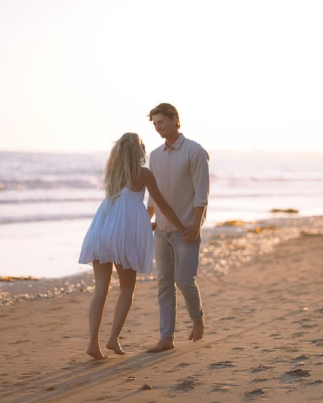 A couple walks barefoot along the beach at sunset. The woman wears a white dress, and the man wears a light shirt and pants. The ocean and soft sunlight create a warm, romantic atmosphere.