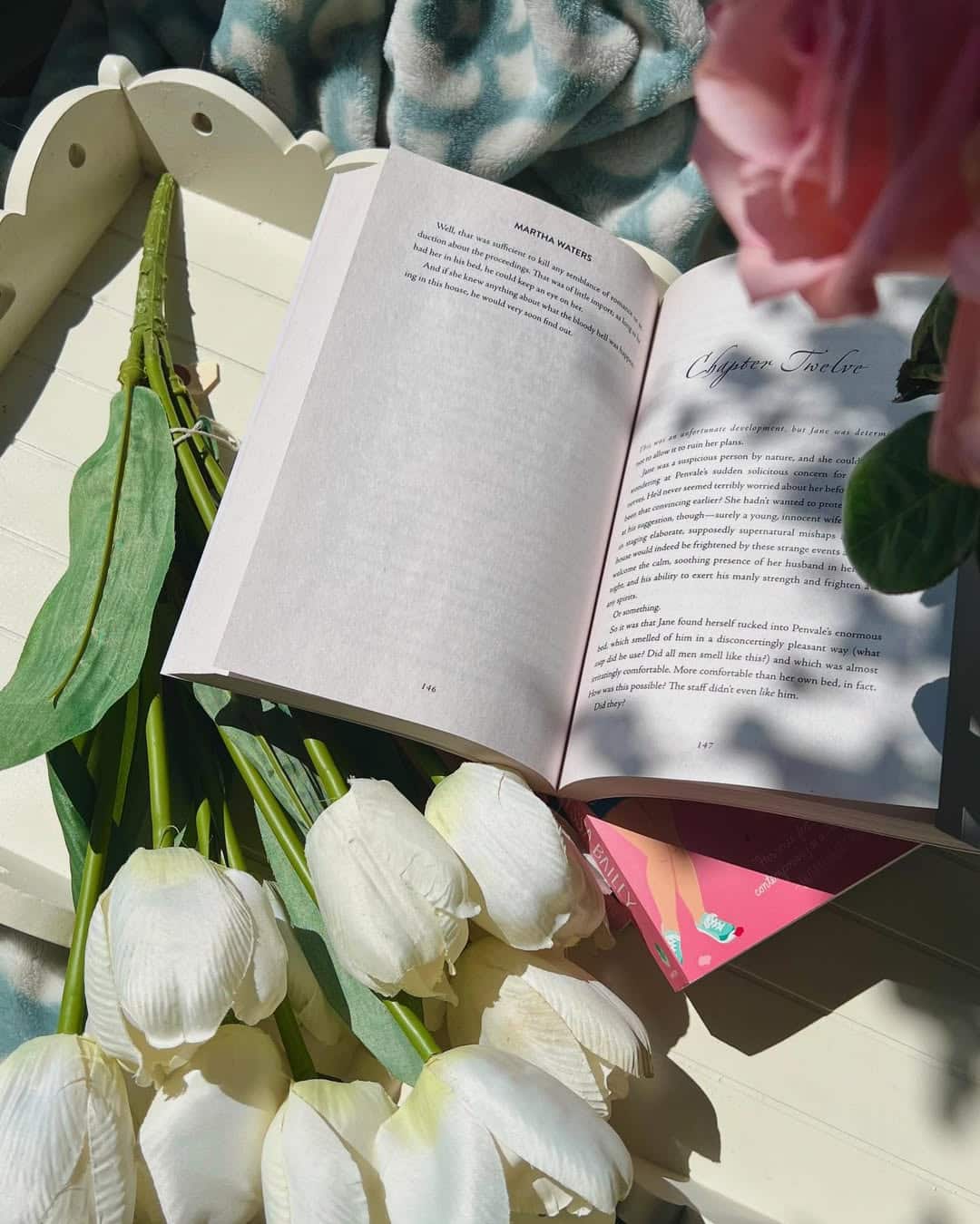 An open book from my TBR rests on a white tray beside a bouquet of white tulips, with sunlight streaming in and pink flowers partially visible in the corner. A soft blanket adds coziness to this serene spring reads scene.