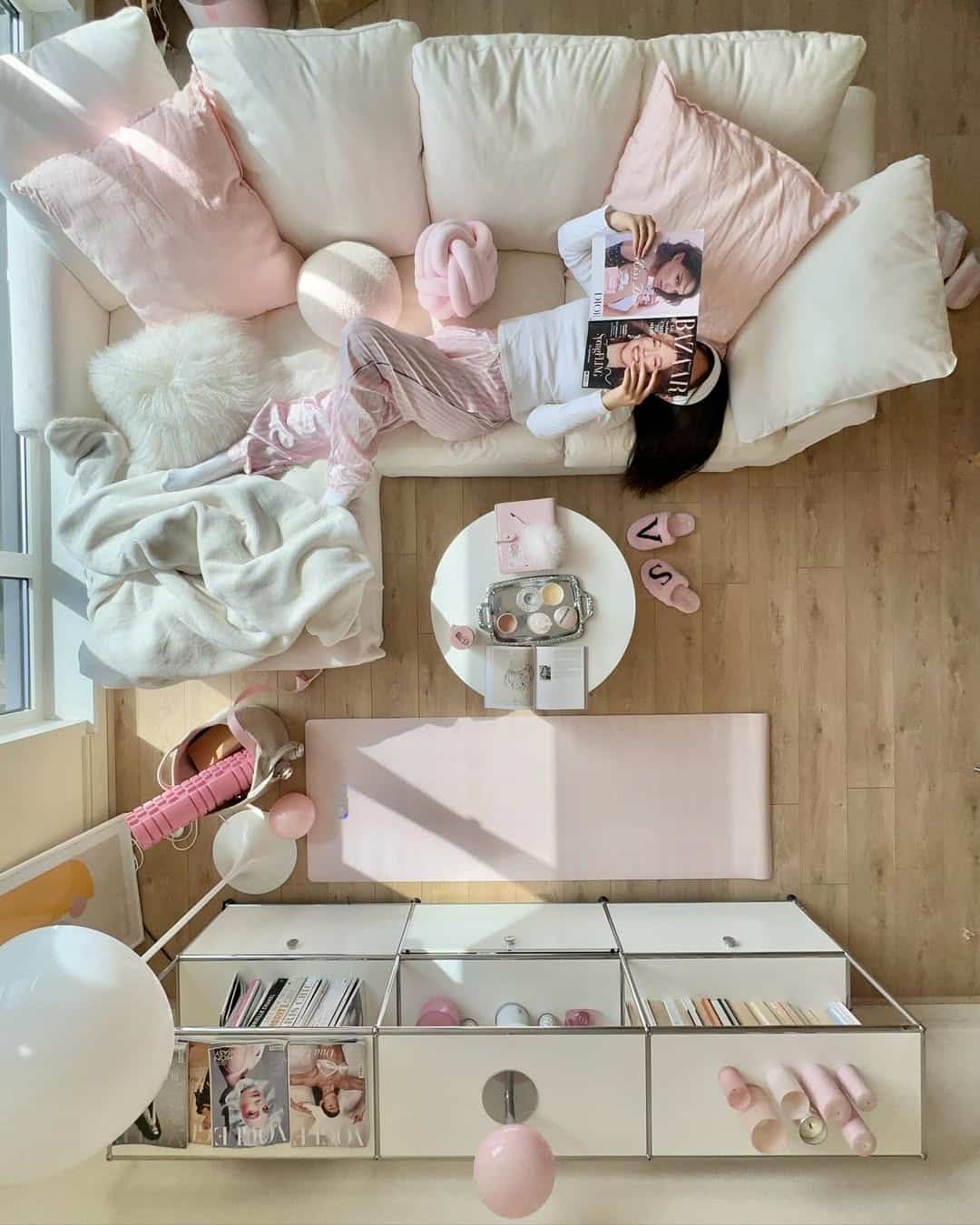 A woman in pink striped pajamas lounges on a white sofa with pink pillows, reading a magazine. A tidy living room with a white coffee table, makeup, a pink yoga mat, and pink and white decor is seen from above.