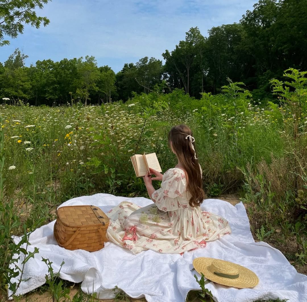 A woman in a floral dress sits on a blanket in a wildflower field, reading a book. A straw hat and picnic basket are beside her, with green trees and a blue, partly cloudy sky in the background.
