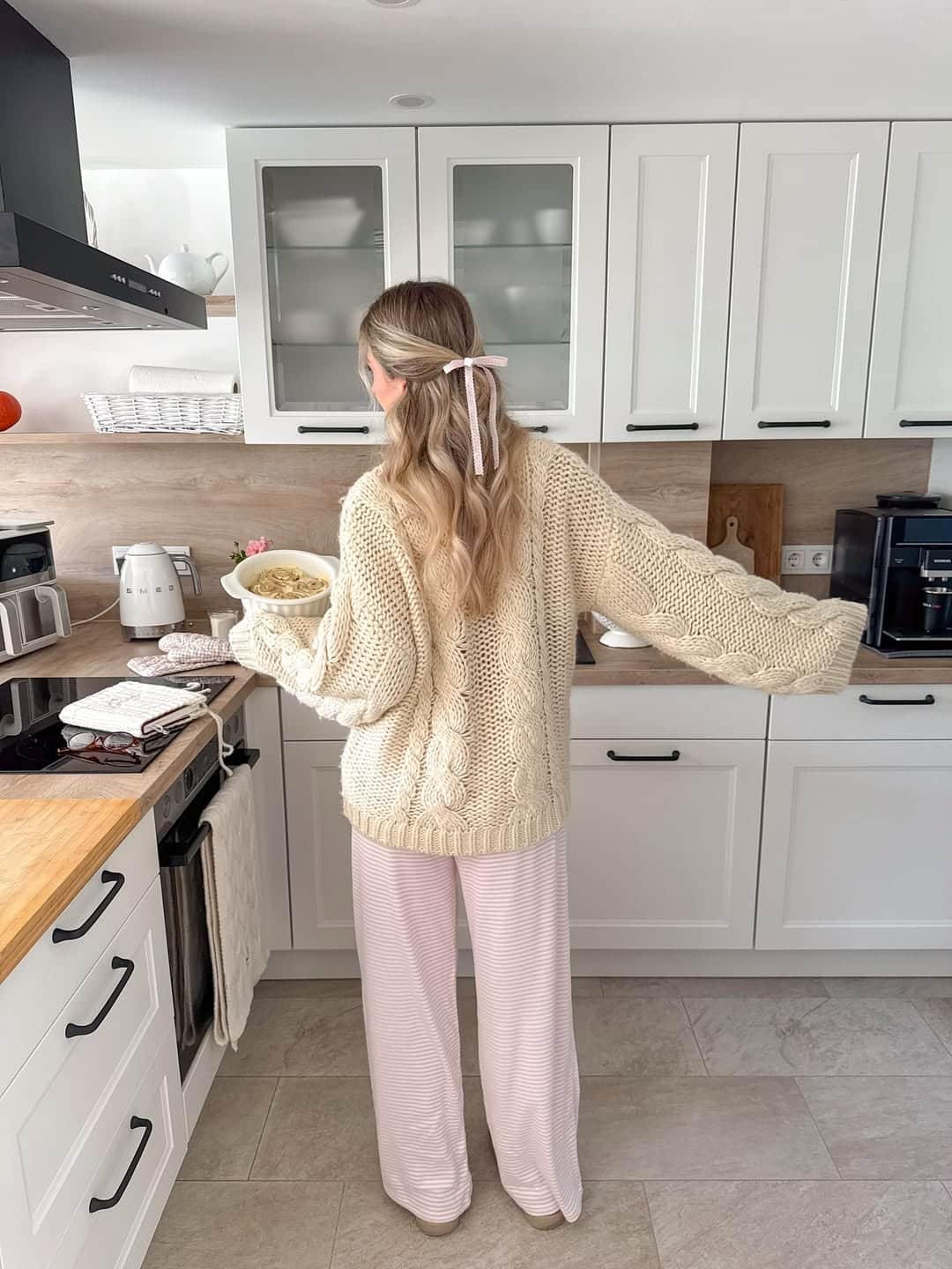 A woman with long blonde hair, tied with a pink ribbon, stands in a modern white kitchen wearing a chunky knit sweater and pink pajama pants, holding a bowl of food while facing away from the camera.