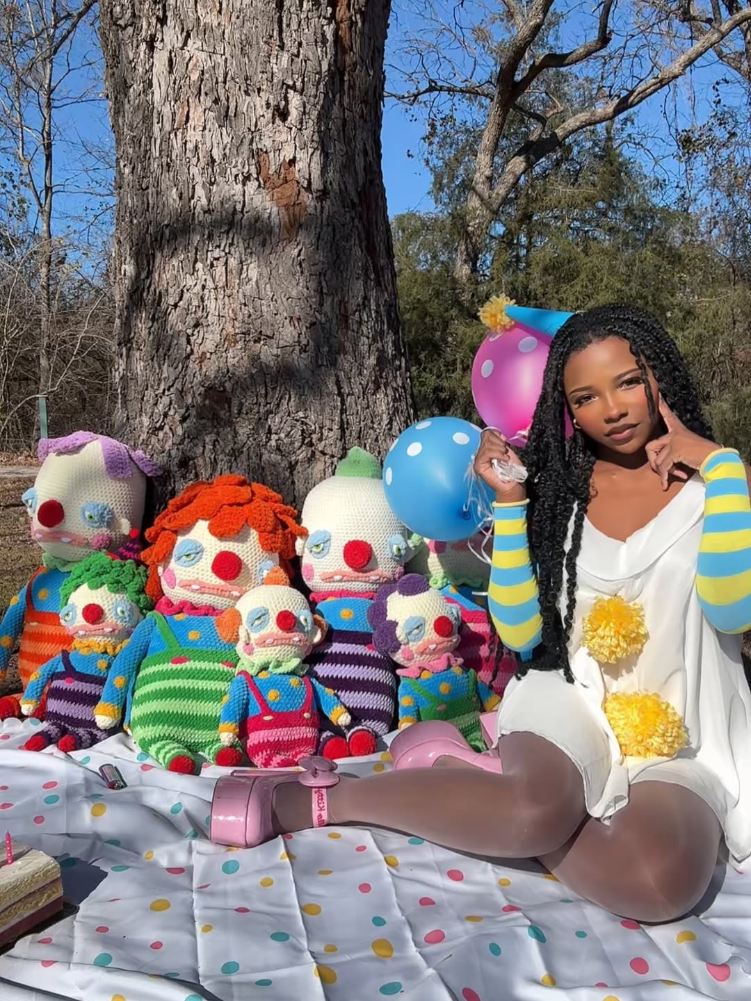 A woman in a white dress with yellow pompoms sits on a polka-dot blanket outdoors, holding a blue balloon, surrounded by large, colorful knitted clown dolls. A tree and greenery are in the background.