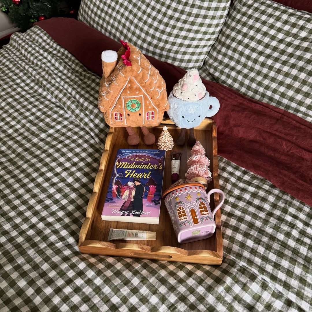 A wooden tray on a green plaid bedspread holds a Christmas mug, the book Midwinters Heart, plush gingerbread house and cupcake, a candle, and holiday-themed decorations—a cozy snow day starter pack.