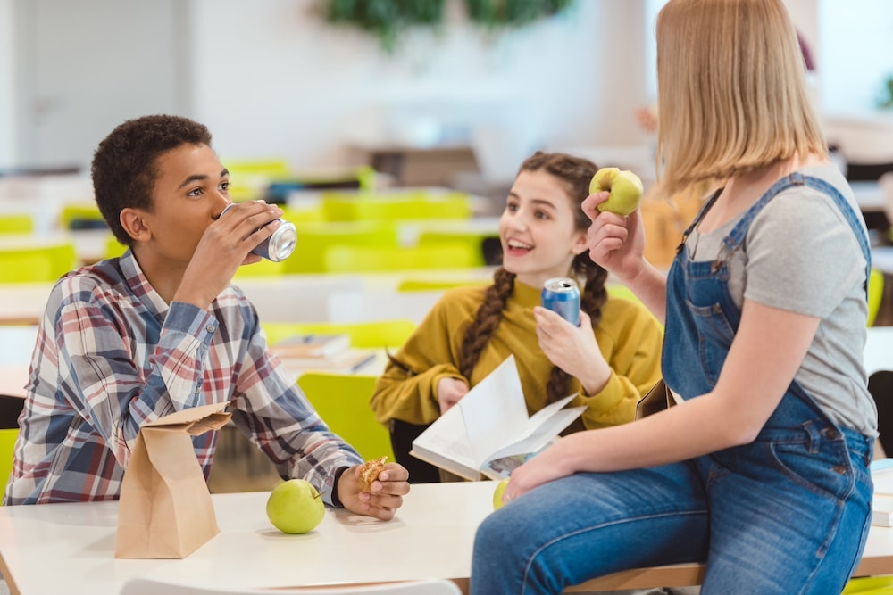 Three students sit at a cafeteria table. One boy drinks from a can, while a girl reading a book smiles. A third girl with an apple sits on the table, chatting about awkward moments. A brown paper lunch bag rests nearby.