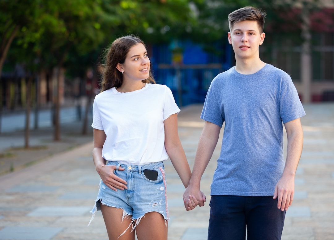 A young couple walks hand in hand outdoors. The woman, wearing a white t-shirt and denim shorts, smiles at the man after an awkward kiss. Trees and buildings are visible in the background.