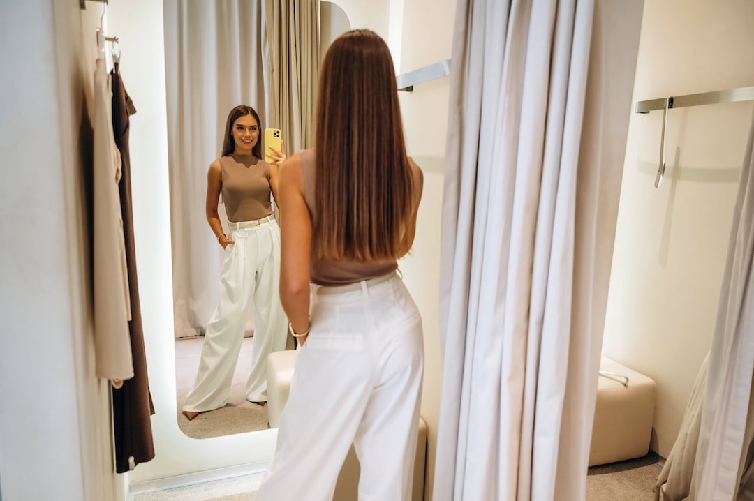 A woman with long straight hair, wearing a beige sleeveless top and white high-waisted pants, stands in a fitting room, pausing for a moment as she notices an awkward moment with her outfit while gazing at her reflection in the full-length mirror.
