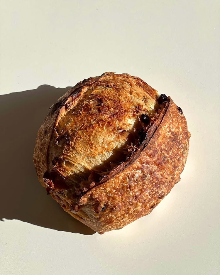 A round loaf of golden-brown artisan bread with a crusty exterior, highlighted by sunlight casting a shadow on a pale surface.