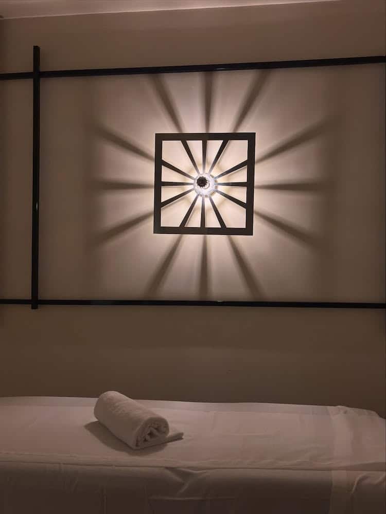 A neatly rolled white towel rests on a massage table beneath a wall-mounted light fixture that casts geometric shadows on the wall, creating a relaxing spa-like atmosphere.