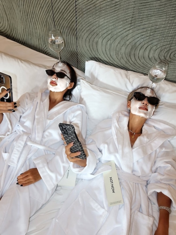 Two people in white robes and sunglasses relax on a bed with white pillows, wearing facial masks. They hold smartphones, balance wine glasses on their heads, and lounge near a skincare box—perfect indoor activities for freezing weather.