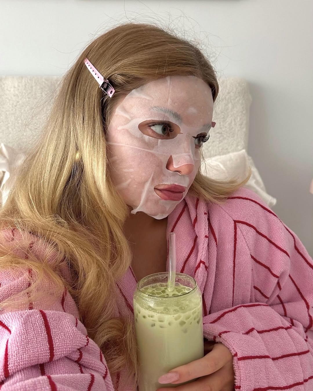 A woman with long blonde hair, wearing a pink striped robe and a pink hair clip, enjoys an at home spa day with a white sheet mask on her face and holds a glass of green iced drink with a straw.