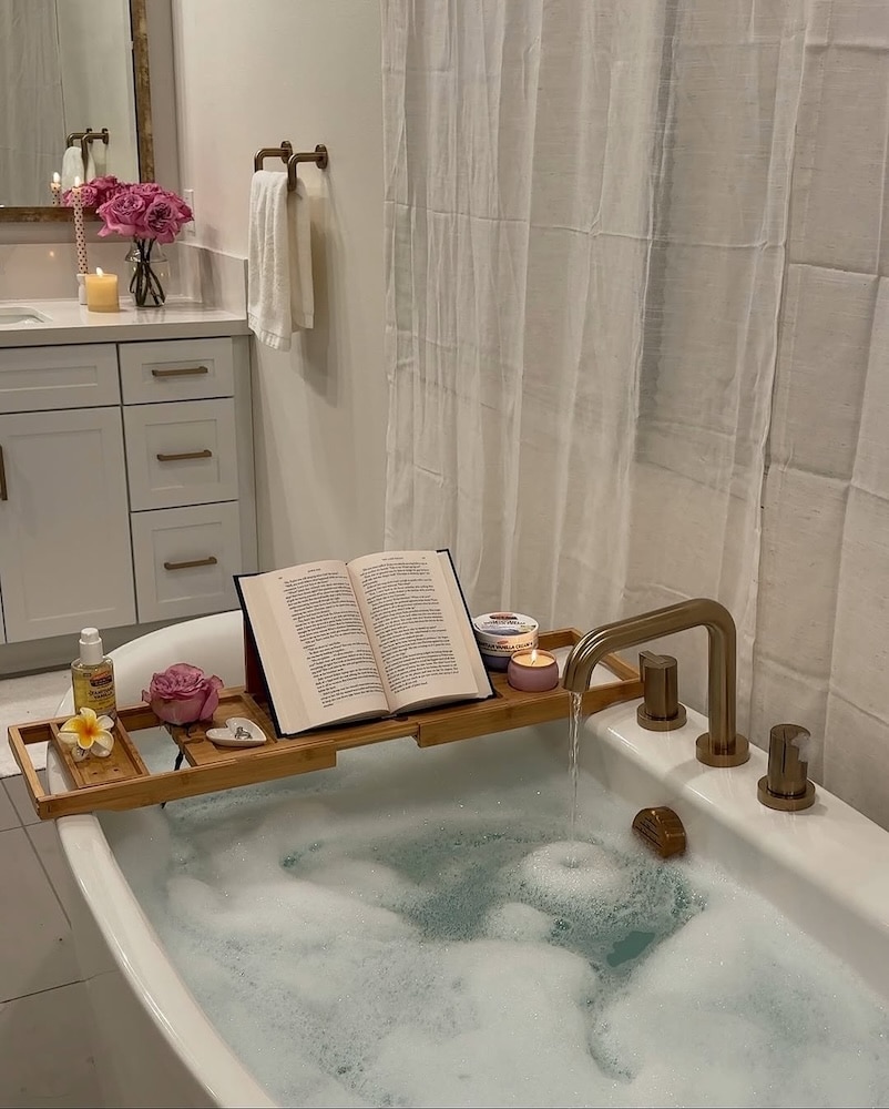 A relaxing at-home spa scene with bubbles, a wooden tray holding an open book, a candle, flowers, and bath essentials; water flows from a gold faucet, with a vanity and pink flowers in the background—a perfect spa day retreat.