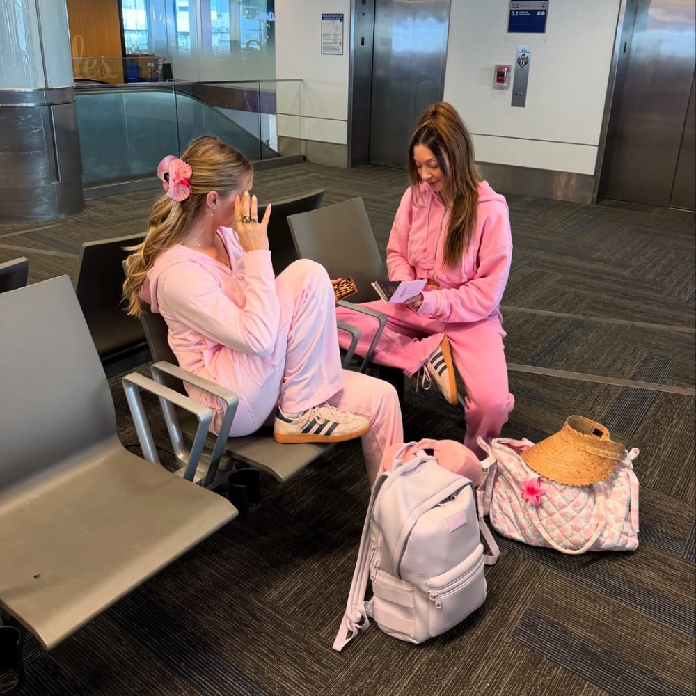 Two women in matching pink spring break outfits sit and chat in an airport waiting area. With light pink backpacks and a straw hat nearby, both are smiling and relaxed while looking at a phone.