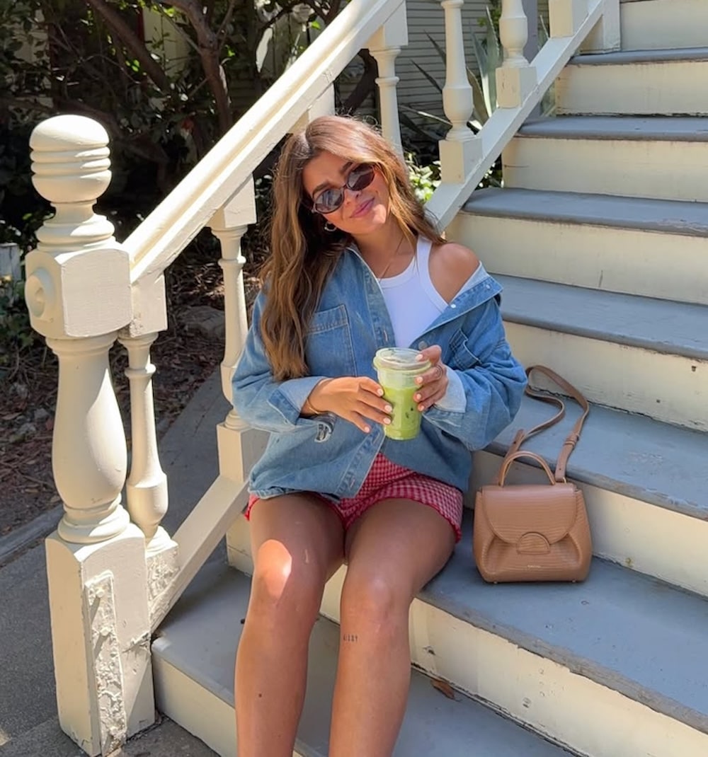 A woman wearing sunglasses, a white top, denim jacket, and red shorts sits on outdoor steps holding a green iced drink. Her look is perfect for spring break fashion, with a tan handbag by her side as she smiles at the camera in the sunlight.