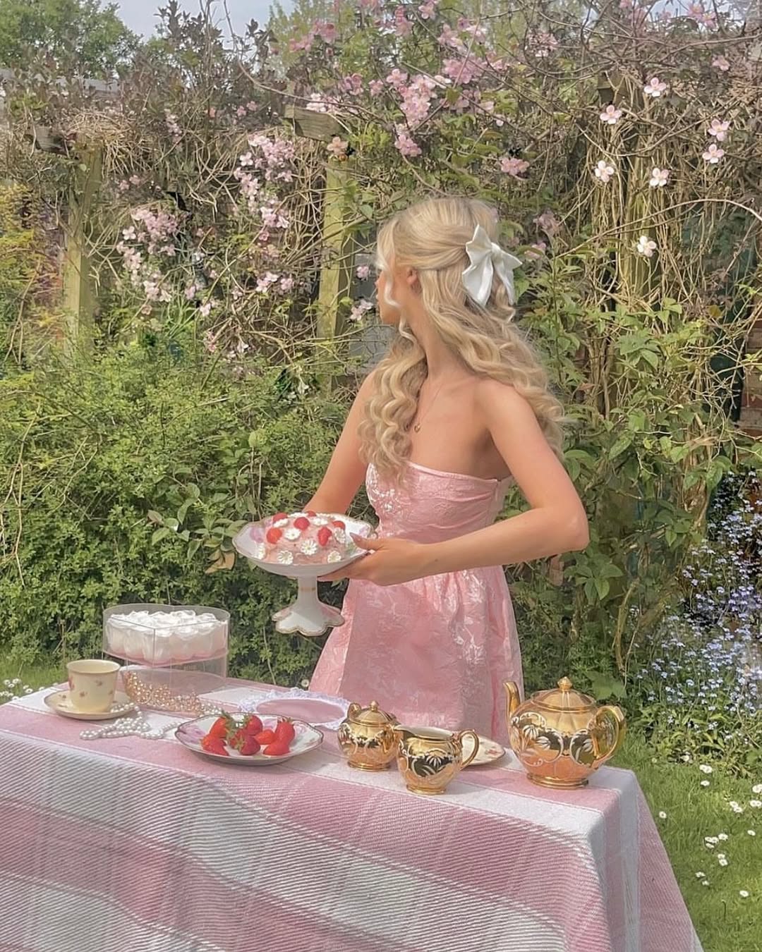 A woman in a pink dress with a white bow in her hair stands at an outdoor table set for a spring brunch, holding a cake. The table has teacups, a teapot, desserts, and sits amidst blooming flowers and greenery.