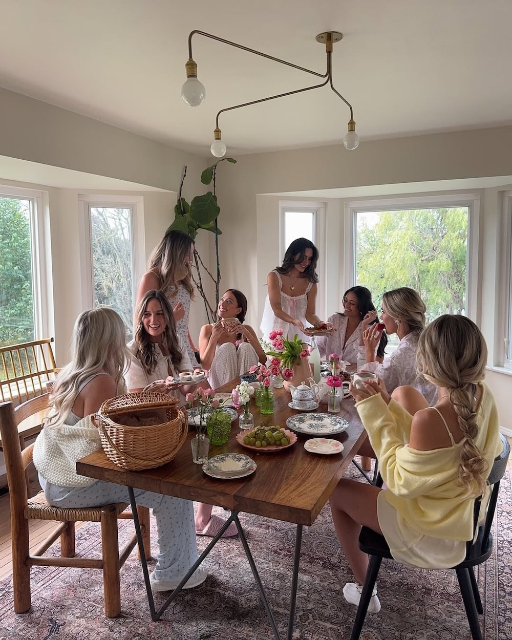 Seven women in light, pastel clothing gather around a wooden table set for a Spring brunch, with flowers, plates, and snacks in a sunlit room. This cheerful squad is smiling, talking, and enjoying a cozy meal together.