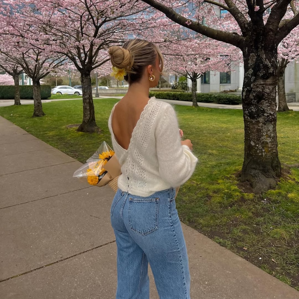 A woman with her hair in a bun, wearing a white sweater and blue jeans—one of the classic spring break outfits—holds a bouquet of yellow flowers while walking on a sidewalk lined with blooming cherry blossom trees.