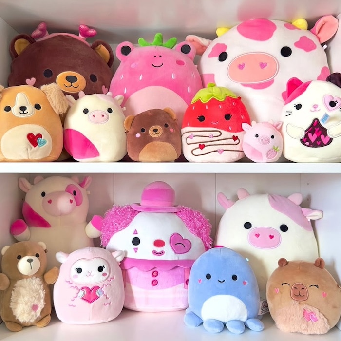 A collection of colorful plush toys, including bears, cows, pigs, a clown, an octopus, a strawberry, and other cute animals, arranged neatly on two white shelves.