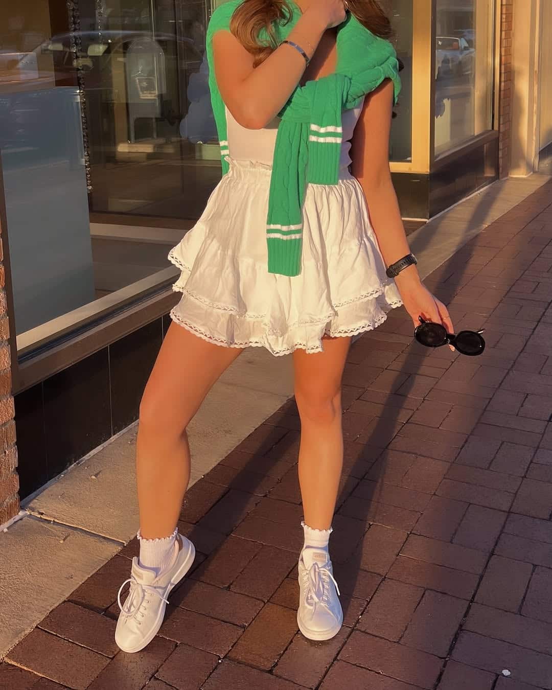 A person stands on a brick sidewalk in a white ruffled dress, white sneakers, and a green sweater draped over their shoulders—making this sunlit, stylish look perfect inspiration for festive outfits or St. Patty's outfits. Black sunglasses complete the ensemble.