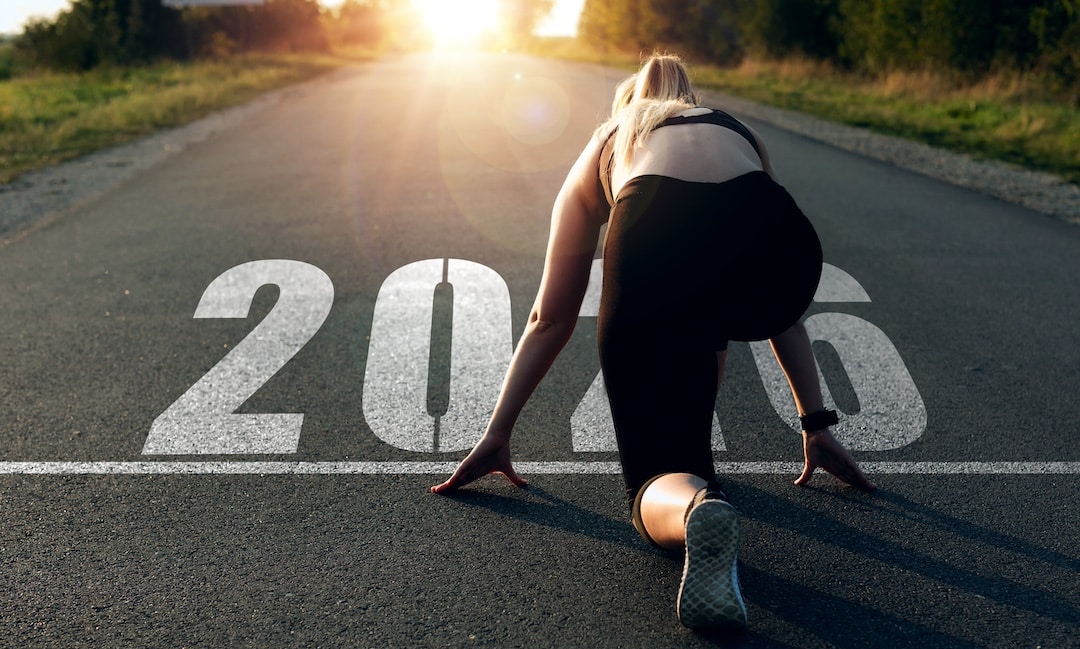 A woman in athletic wear is in a starting position on a road with 2026 painted on the ground, facing a bright sunrise—embracing a 2026 mantra of fresh starts and new goals for the year.