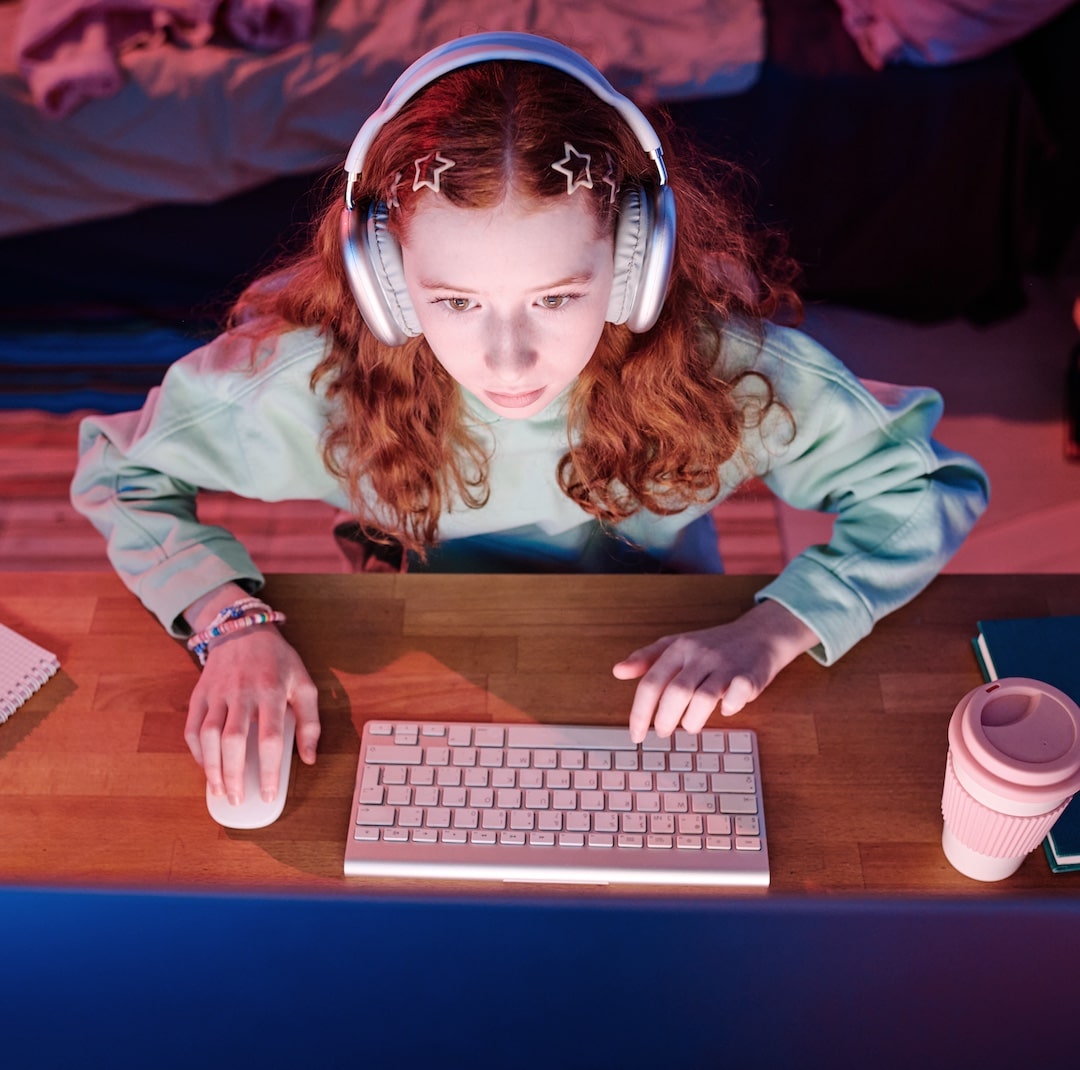 A girl with long red hair, wearing headphones and a green sweatshirt, sits at her desk deep into a gaming sesh. With a reusable cup and notebook beside her, she looks focused on the screen, ready to team up with her virtual bestie.