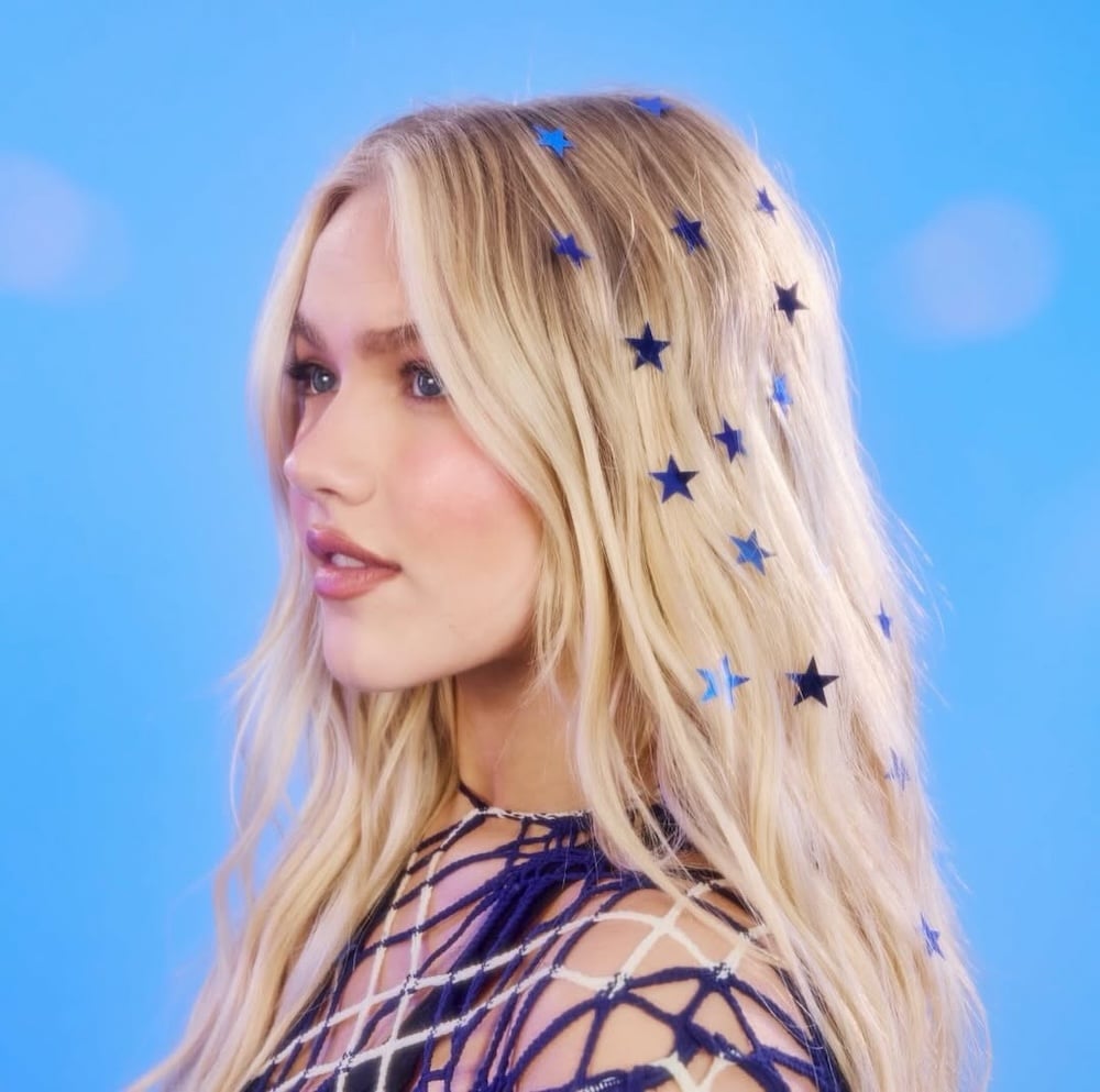 A woman with long blonde hair decorated with blue star-shaped confetti channels stardust vibes as she looks to the side against a bright blue background—her soft, natural makeup and netted top evoke the artistry of Freya Skye’s latest EP.