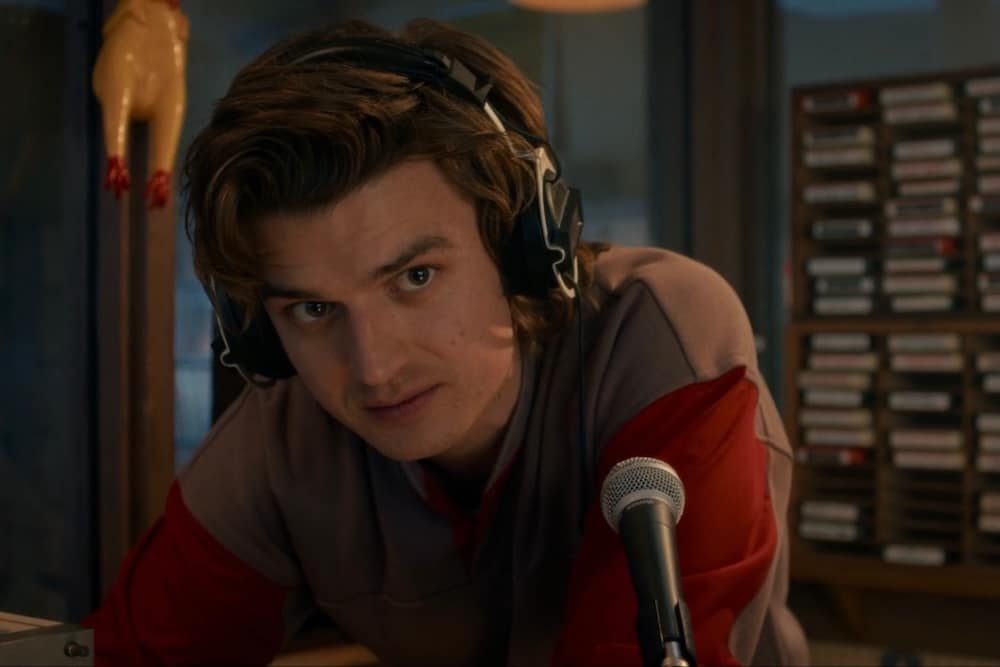 A young man with wavy brown hair, resembling a Zodiac character, wears large headphones and leans toward a microphone in a recording studio, with cassette tapes and a rubber chicken visible in the background.
