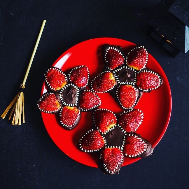 A red plate holds three flower-shaped chocolate treats, each made with five strawberry halves and decorated with white sprinkles, on a dark background. A small broomstick and black lantern are nearby.