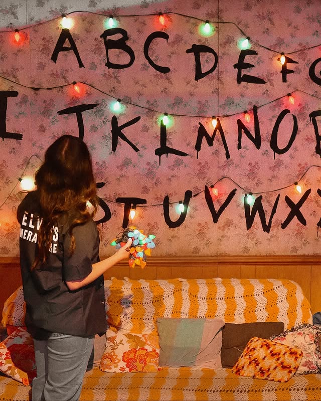 A person, likely the host of a Stranger Things finale watch party, stands in front of a wall decorated with the alphabet and colorful string lights. Holding more lights and wearing a black top, they face away from the camera.
