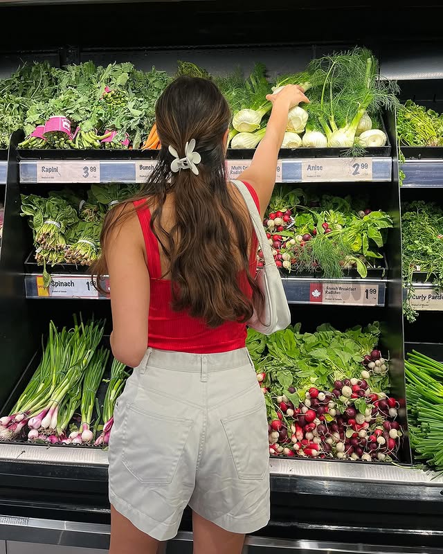 A woman with long brown hair, wearing a red tank top and beige shorts, stands in front of a grocery store vegetable display, reaching up to pick produce. Fresh greens and radishes are visible on the shelves.