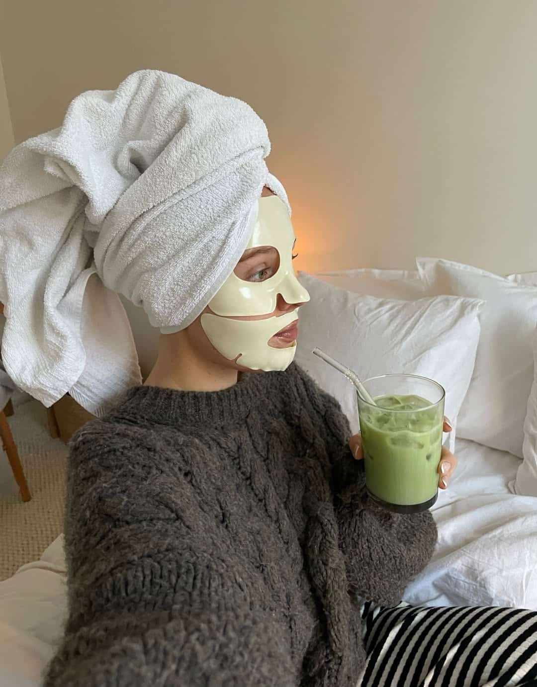 A person wearing a face mask and towel wrapped around their head sits on a bed, sipping an iced green drink—embracing a cozy sweater and striped pants for the perfect Sunday Reset Routine in a relaxed, self-care atmosphere.