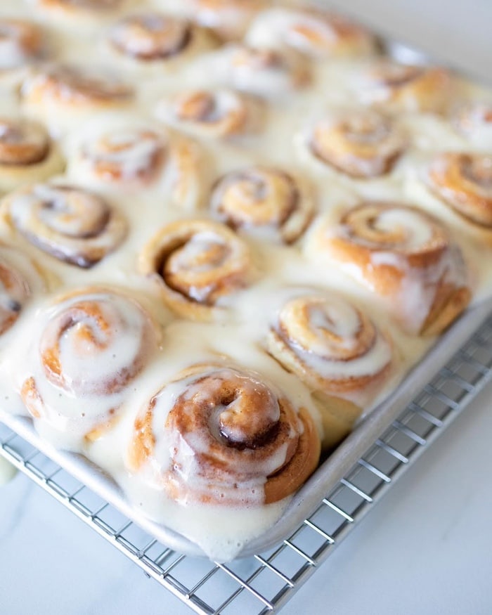A tray of freshly baked cinnamon rolls covered in creamy white icing, resting on a metal cooling rack. The rolls are golden brown and closely packed together.