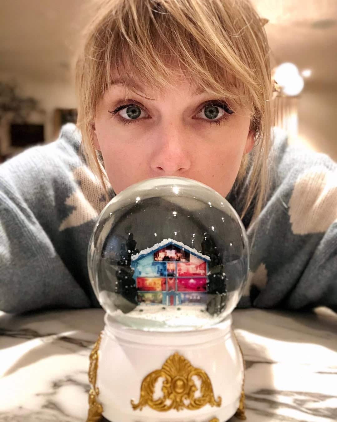 A woman with blonde hair rests her chin on a table, looking closely at a decorative snow globe featuring a festive house scene inside—perfect for those seeking Swiftmas ideas to celebrate Swiftmas in a cozy, magical way.