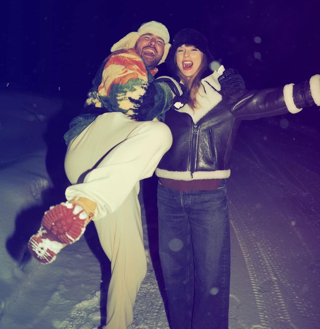 Two people pose playfully on a snowy road at night, celebrating Swiftmas. One in light pants and a colorful jacket lifts their leg high, while the other, in a dark jacket and hat, smiles with arms outstretched amid the snow-covered ground.