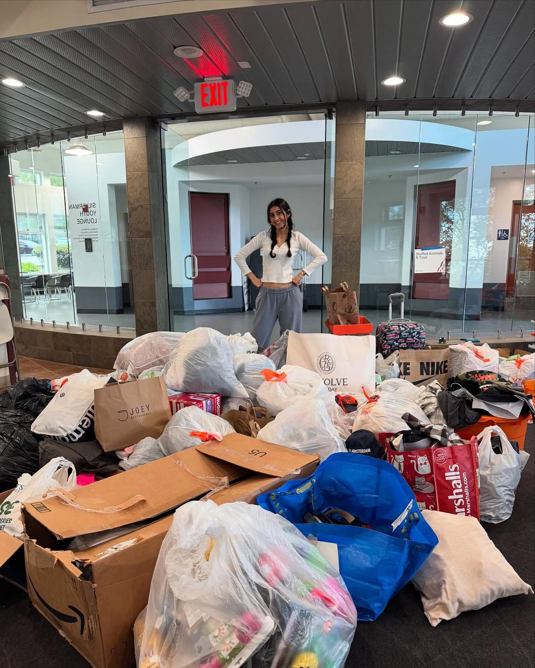 A woman stands smiling behind a large pile of assorted bags and cardboard boxes filled with donated items inside a modern building lobby.